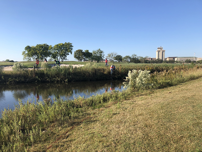 A scenic view of individuals working along a grassy bank by a calm waterway, with a building in the background under a clear blue sky.